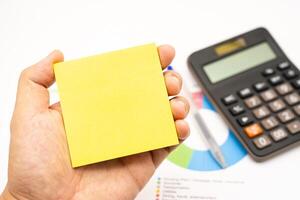 Hand holding Blank Sticky note with calculator background. Financial data analyzing. Working on desk with using calculator, finance accounting. Copy space. photo