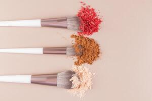 Three professional fluffy brushes with different samples of loose dry makeup textures on a beige background. top view. A copy of the space. photo