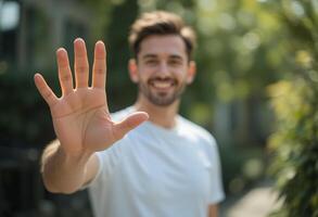 Professional Man Smiling in White T-Shirt Making Hand Gesture, Hello, Stop or High-Five Signal Against Blurred Garden Background photo