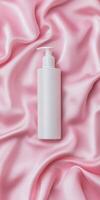 Minimalist flat lay of glossy white bottle on soft pink satin background for skincare design photo