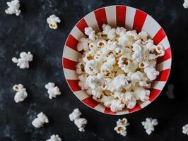 Salted popcorn in striped bowl overhead spill on dark surface for snack themed designs photo