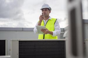 Asian maintenance engineer works on the roof of factory. contractor inspect compressor system and plans installation of air condition systems in construction. technology, online, mobile application photo
