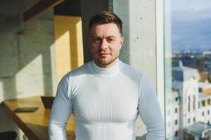 A young handsome man in a white sweater is standing in a spacious bright office. The concept of a modern successful person. Young smiling guy in open office space photo