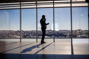 Rear view of a businessman looking out of a large window overlooking the city. He has a phone in his hands. Horizontal view. photo