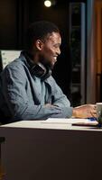Vertical African american analyst works on accounting analysis while he is dancing, feeling the rhythm and checks financial balance. Remote worker listening to favorite groovy music on headphones. Camera A. video