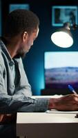 Vertical African American freelancer working from home writing notes and researching data driven topics. Focused on productivity and planning tasks in a modern apartment for remote work success. Camera A. video