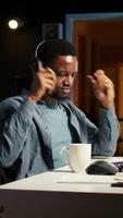 Vertical Young casual guy listening to a podcast while he works on freelance job at home, enjoys multitasking with headphones. Man solving tasks and paying attention to webinar discussion. Camera A. video