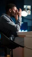 Vertical Young black employee with a migraine yawning at home workstation, examining online reports to solve new management tasks. African american guy being tired and stressed of project deadline. Camera A. video
