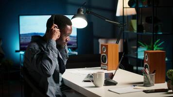 African american specialist closing the monthly balance with a show on headset, working remotely while he listens to his favorite podcast. Managing expenses and insurances from home. Camera B. video
