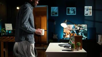 Self employed black man sitting at the desk organizing his schedule, reviewing online data notes. Guy with a coffee cup browsing the internet for research, remote work in a modern loft. Camera A. video