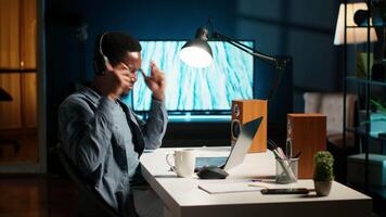 Young casual guy listening to a podcast while he works on freelance job at home, enjoys multitasking with headphones. Man solving tasks and paying attention to webinar discussion. Camera B. video