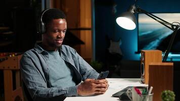African american person takes a break from work to answer texts at home, using his smartphone to scroll social media apps. Young guy enjoying remote job at home, listens to music on phone. Camera B. video