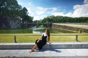 Woman Sitting by Moat with Bridge and Greenery in Tokyo on a Sunny Day photo