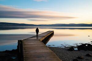 Solitary Figure Walking on a Wooden Pier Over a Calm Lake at Sunset with Reflective Water photo
