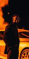 Stylish man in a sleek black outfit posing by an orange sports car under dramatic lighting for cinematic mobile wallpaper photo