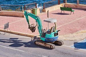 Compact excavator digging holes for palm tree planting as part of an urban landscaping project near a waterfront. Xghajra, Malta-16.06.2020 photo
