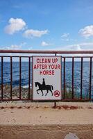 Sign instructing horse riders to clean up after their horses is mounted on rusted metal railing along scenic coastal pathway photo
