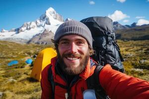 Smiling backpacker taking selfie with snowy mountain backdrop and camping tents in scenic outdoor adventure setting photo