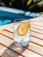 Glass of refreshing ice water with a lemon slice on a sunny patio table near a pool photo