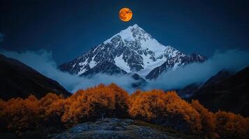 Majestic Snow Capped Mountain Peak at Night with a Full Orange Moon and Autumnal Trees photo