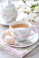 Delicate tea cup with floral design accompanied by pink macarons on a patterned napkin and flowering branches in a serene setting photo