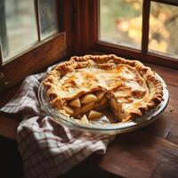 A freshly baked apple pie with a golden, flaky crust sits on a wooden windowsill. A slice is missing, revealing tender apple filling. Soft daylight filters through the window, casting warm tones photo