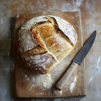 A top-down view of a whole rustic loaf of bread on a wooden cutting board. The crusty surface and rich texture stand out, with a knife placed beside it, highlighting the simplicity of homemade baking photo
