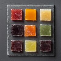 A minimalist arrangement of fruit jellies in a square grid on a dark slate plate. The candies include shades of red, orange, yellow, green, and dark purple, with a sugar-coated texture photo