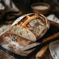 A half loaf of rustic bread with a crisp crust sits on a wooden board. A knife lies beside it, ready to slice. A sprig of rosemary adds a fresh aromatic touch, enhancing the artisanal baking feel photo
