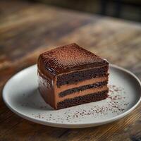 A triple-layer chocolate cake slice covered in glossy ganache and sprinkled with cocoa powder. Served on a white plate with crumbs scattered around, resting on a rustic wooden table photo