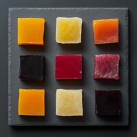 Nine square fruit jelly candies of different colors displayed on a dark slate surface. The candies appear soft and slightly glossy, with hues ranging from bright yellow to deep red and dark purple photo