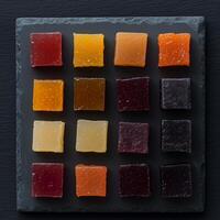A selection of fruit jelly candies arranged in a square grid on a black slate plate. The candies vary in colors, including red, yellow, orange, and dark purple, with a slightly translucent texture photo