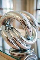 A shiny metal mobius strip knot on a glass table, in a room with a window and curtains in the background photo