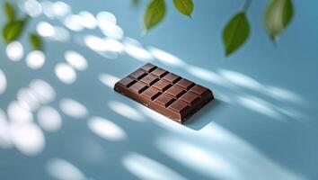 Dark chocolate bar resting on a blue surface with shadow patterns created by leaves in sunlight photo