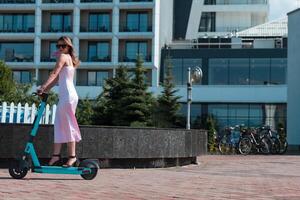Elegant woman in a pink dress and sunglasses riding an electric scooter through the sunny city streets, embracing the freedom and style of a perfect urban adventure. photo