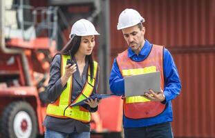 Female supervisor in safety gear using clipboard checklist and foreman worker in hardhat holding laptop for control loading containers box from cargo photo