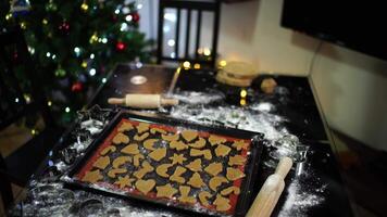 Cookies cut from ginger dough lie on a baking sheet on the table near the Christmas tree in the room video