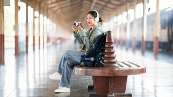 An asian woman is taking a photo with her camera while sitting on the bench in the train station.