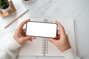 Close up of the pair of hand holding smartphone horizontally over an open notebook on marble table. photo