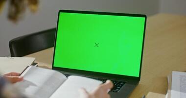 A young man of European appearance is leafing through a book while sitting in front of an open laptop. Filming from behind, copy space, chroma key, focus on green laptop screen video