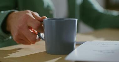 The man takes the mug from the table next to the office papers and takes a sip and puts it back. Coffee break during work, close-up of a mug, unidentified person video