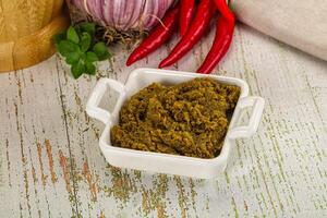 A bowl of green curry paste on a table next to a knife and a pepper photo