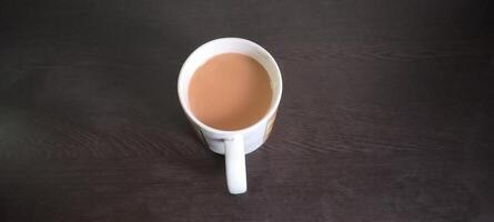 A white mug filled with creamy, milky tea sits on a dark wooden table. The smooth, rich color of the tea contrasts with the deep tones of the wood, creating a cozy and inviting scene. photo