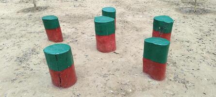 Six wooden stumps painted green and red stand on dry ground, arranged in a playful pattern. Surrounded by scattered leaves, the setting gives a rustic yet vibrant look, possibly part of a park. photo