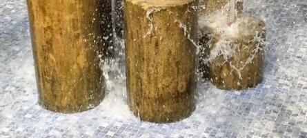 Water cascades over cylindrical wooden stumps in a fountain, creating dynamic splashes. The stumps stand on a mosaic-tiled surface, enhancing the rustic-meets-modern aesthetic of the water feature. photo