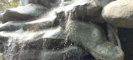 Water gracefully flows down an artificial rock structure, mimicking a natural waterfall. The glistening droplets catch the sunlight, while fallen leaves add a touch of nature to the scene. photo