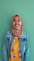 A woman wearing a hijab smiles while standing in front of a green wall photo