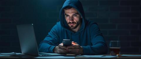 Man In Hoodie Using Smartphone With Laptop on Desk photo