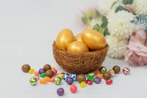 Golden Easter eggs in bird nest and bright pink and white flowers on white table on white wall background photo