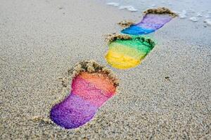 Rainbow colored footprints leading forward on sandy beach, symbolizing progress in rights activism during pride month photo